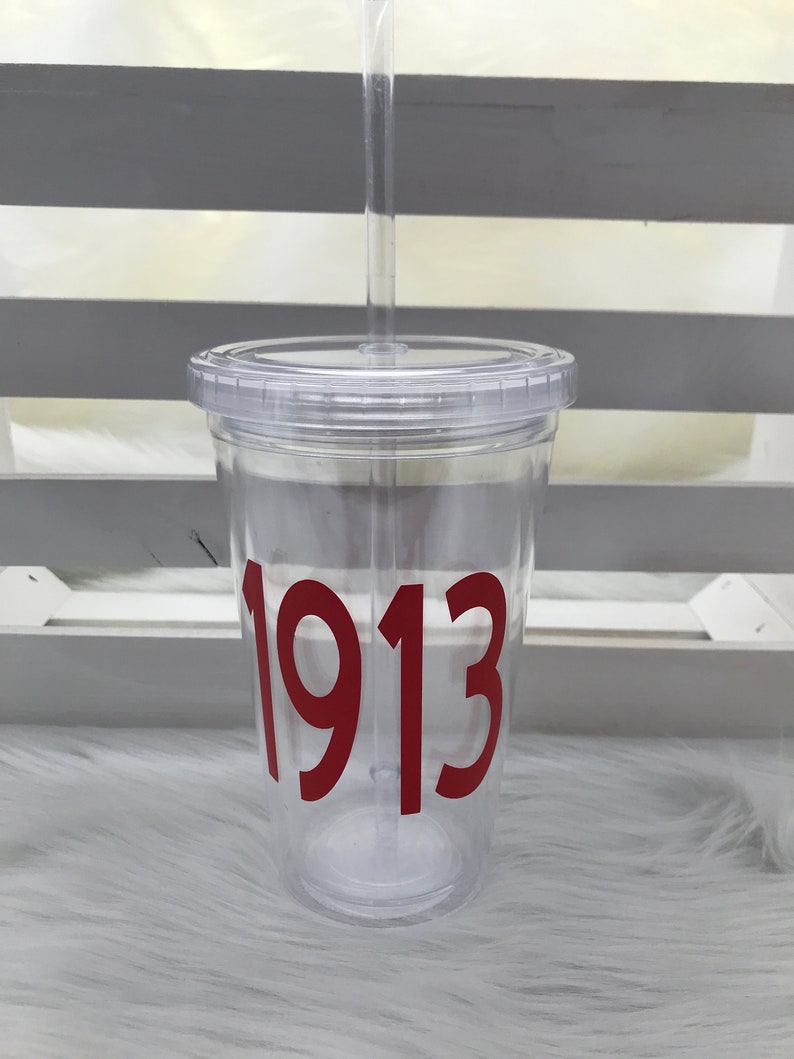 May include: Clear plastic tumbler with a lid and straw. The tumbler features the number "1913" in a bold, red font. The cup is designed for cold beverages and is shown on a white, fluffy surface.