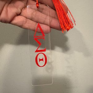 May include: A clear acrylic bookmark with red Greek letters and a matching red tassel. The bookmark features the Greek letters Delta, Sigma, and Theta. The tassel is made of multiple strands of red thread.