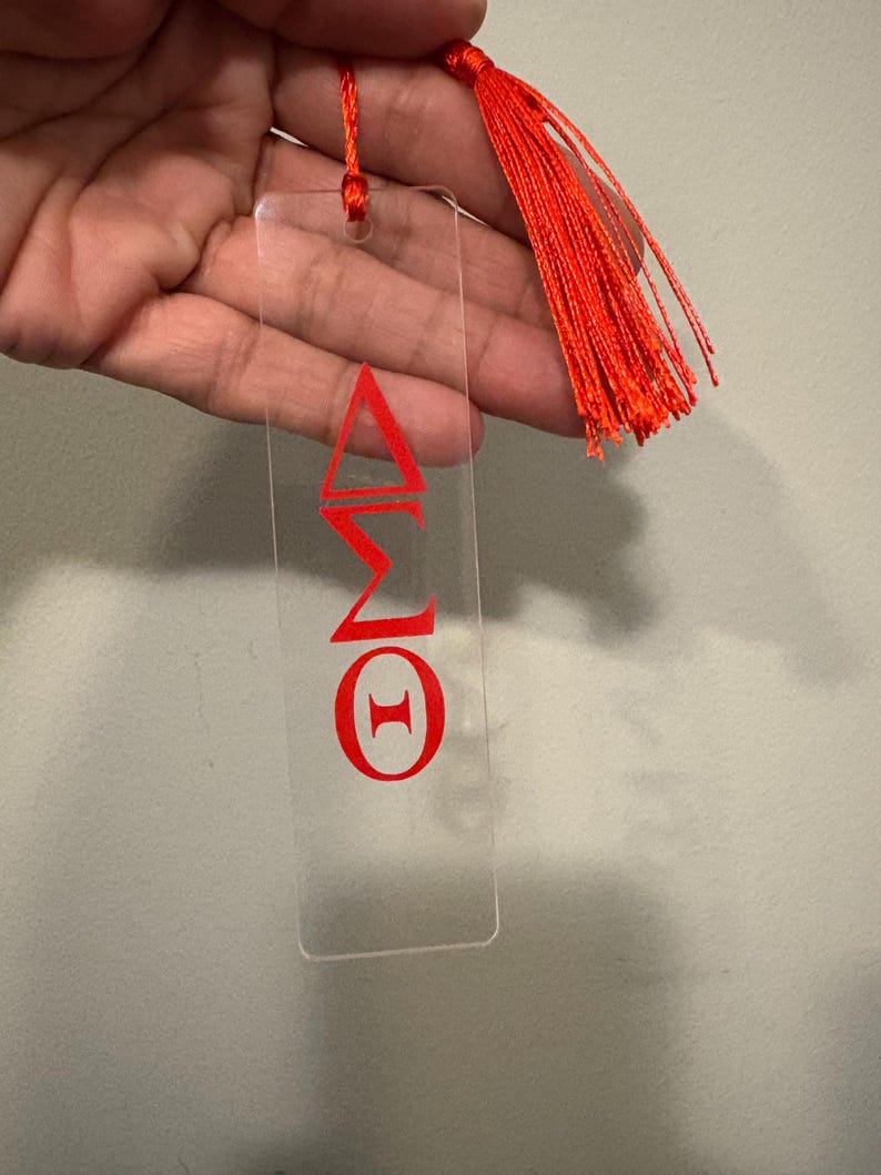 May include: A clear acrylic bookmark with red Greek letters and a matching red tassel. The bookmark features the Greek letters Delta, Sigma, and Theta. The tassel is made of multiple strands of red thread.