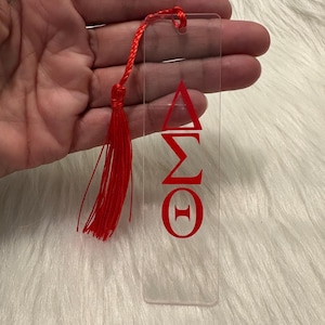 May include: Clear acrylic bookmark with red Greek letters and a red tassel. The bookmark features the Greek letters Delta, Sigma, Omega, and Theta. The tassel is attached to the top with a red cord.