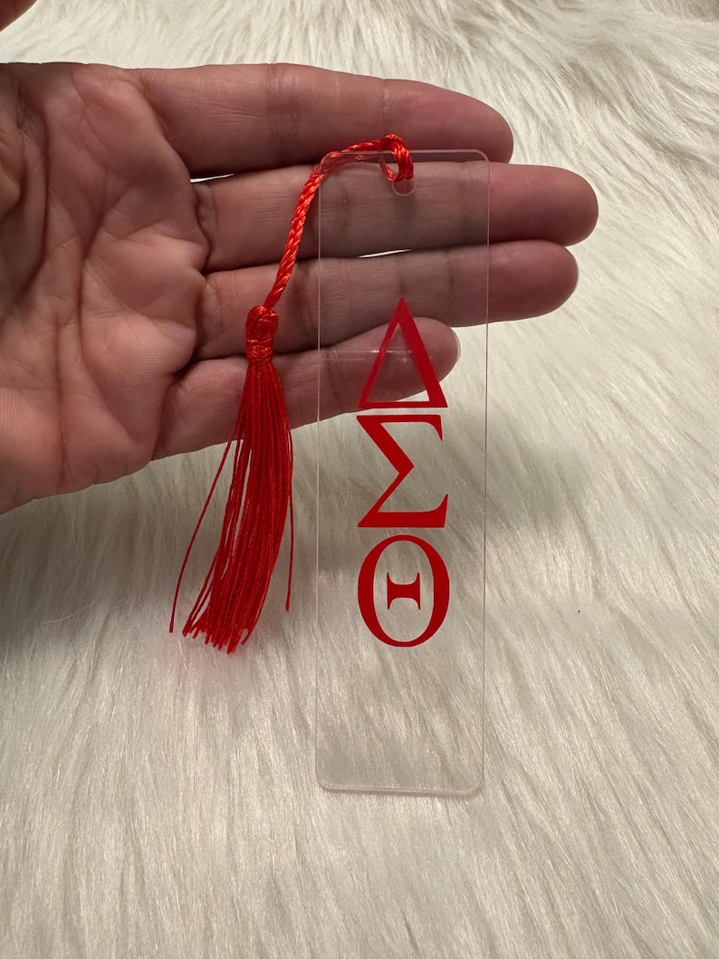 May include: Clear acrylic bookmark with red Greek letters and a red tassel. The bookmark features the Greek letters Delta, Sigma, Omega, and Theta. The tassel is attached to the top with a red cord.
