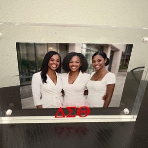 May include: Clear acrylic photo frame with a picture of three women in white outfits. The frame has a black and white border and the Greek letters ΔΣΘ in red. The frame is held together with four silver screws.