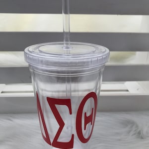 May include: A clear plastic tumbler with a lid and straw. The tumbler features the Greek letters Delta, Sigma, and Theta in red. The tumbler is designed for cold beverages.