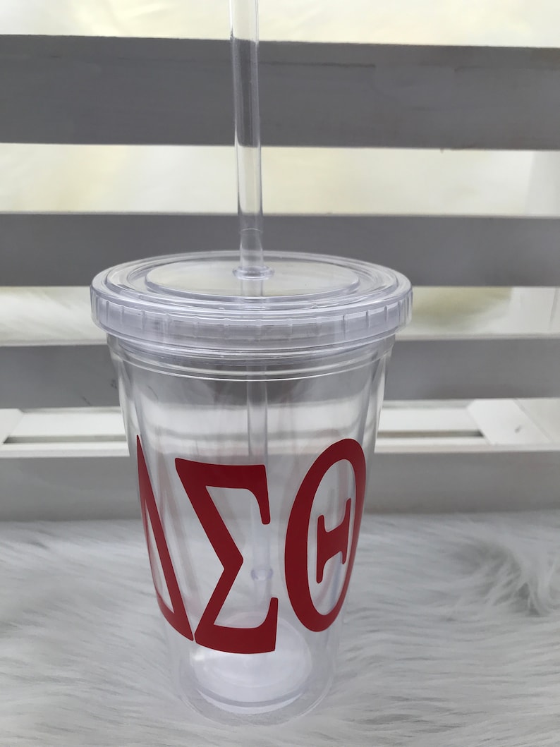 May include: A clear plastic tumbler with a lid and straw. The tumbler features the Greek letters Delta, Sigma, and Theta in red. The tumbler is designed for cold beverages.