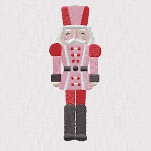 Puede incluir: Imagen bordada de un soldado cascanueces rosa y rojo. El cascanueces tiene bigote y barba blancos, un sombrero rosa y rojo y un cinturón negro. El uniforme del soldado es rosa con detalles rojos y botas negras. La imagen está sobre un fondo blanco.