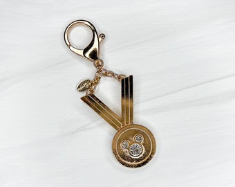 May include: Gold-tone keychain with a Mickey Mouse head charm and a V-shaped design. The charm has a clear rhinestone in the center.