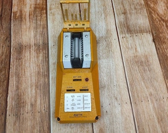 May include: A yellow and silver Geiger counter with a rectangular body and a hinged top. The device has a series of buttons and a display panel with various symbols and text. The device is designed to measure radiation levels.