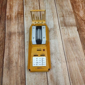 May include: A yellow and silver Geiger counter with a rectangular body and a hinged top. The device has a series of buttons and a display panel with various symbols and text. The device is designed to measure radiation levels.