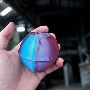May include: A two-tone sphere, divided into blue and purple halves. The object has a textured, layered appearance, and is held in a hand against a blurred background. The top of the sphere has a rough, open surface.