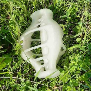 Puede incluir: Una escultura abstracta blanca con un diseño de celosía, apoyada sobre un lecho de hierba verde y trébol. El objeto tiene una parte superior e inferior bulbosa, conectadas por una serie de barras horizontales.