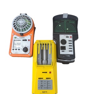 Peut inclure: Trois appareils électroniques vintage. Un appareil orange avec un dessus en forme de ventilateur et un écran, un appareil jaune avec un tube central et un appareil noir avec un écran radar et des boutons.
