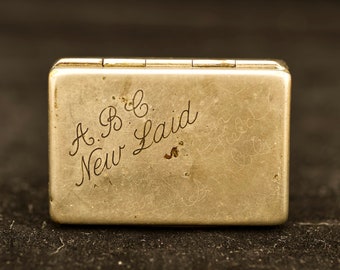 May include: A silver metal cigarette case with the inscription "A.B.C. New Laid" engraved on the front.