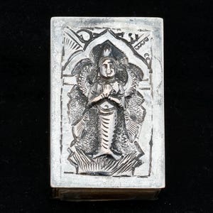 May include: A rectangular silver-colored box with a detailed relief carving of a figure with a crown, wings, and a fish-like tail. The figure is in a praying pose, set within an ornate archway design. The box has a slightly aged appearance.