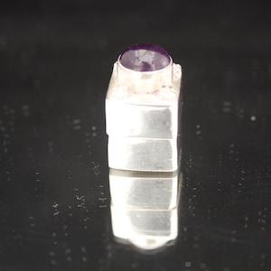 Könnte beinhalten: Eine kleine silberne Schachtel mit einem violetten Edelstein oben. Die Schachtel ist quadratisch und hat einen Scharnierdeckel.