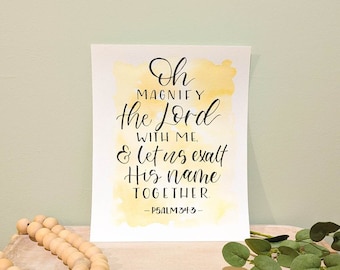 May include: A watercolor print with a yellow background and black calligraphy that reads "Oh Magnify the Lord with me & let us exalt His name together. -Psalm 34:3-"