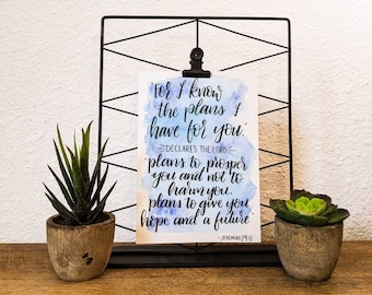 May include: A watercolor print with a blue and white background featuring a quote from Jeremiah 29:11, "For I know the plans I have for you." The quote is written in black calligraphy and is framed in a black metal stand.