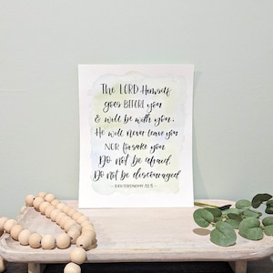 May include: A watercolor print with a white background and a light blue, yellow, and green watercolor wash. The print features a quote from Deuteronomy 31:8: "The LORD Himself goes BEFORE you & will be with you; He will never leave you NOR forsake you Do not be afraid. Do not be discouraged."