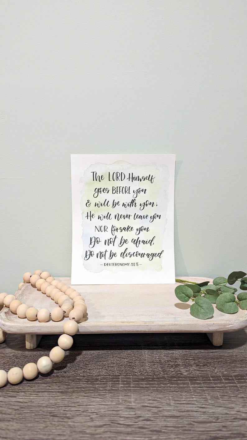 May include: A watercolor print with a white background and a light blue, yellow, and green watercolor wash. The print features a quote from Deuteronomy 31:8: "The LORD Himself goes BEFORE you & will be with you; He will never leave you NOR forsake you Do not be afraid. Do not be discouraged."