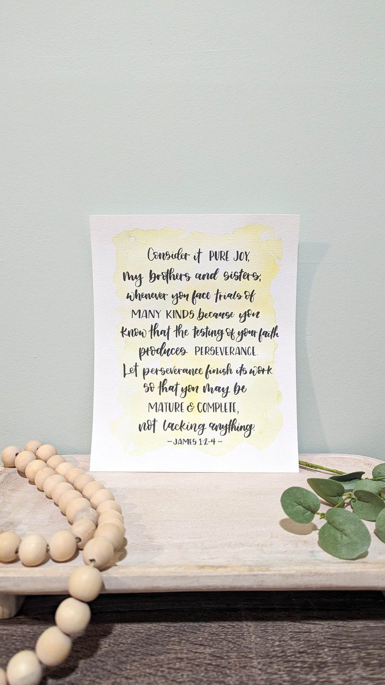 May include: A watercolor print with a yellow background and black text. The text reads "Consider it PURE JOY, my brothers and sisters, whenever you face trials of MANY KINDS because you know that the testing of your faith produces PERSEVERANCE. Let perseverance finish its work so that you may be MATURE & COMPLETE, not lacking anything. -JAMES 1:2-4- "
