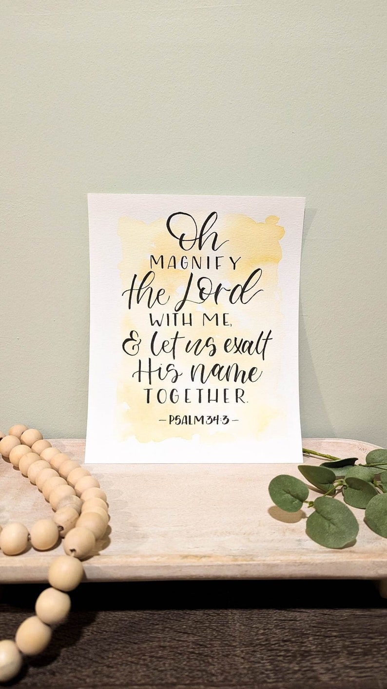 May include: A watercolor print with a yellow background and black calligraphy that reads "Oh magnify the Lord with me, & let us exalt His name together. -Psalm 34:3-"