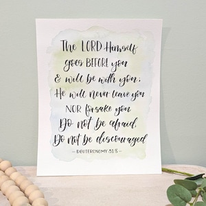 May include: A watercolor print with a light blue, green, and yellow background. The print features a handwritten quote from Deuteronomy 31:8, which reads: "The LORD Himself goes BEFORE you & will be with you; He will never leave you NOR forsake you Do not be afraid Do not be discouraged - DEUTERONOMY 31:8 -"
