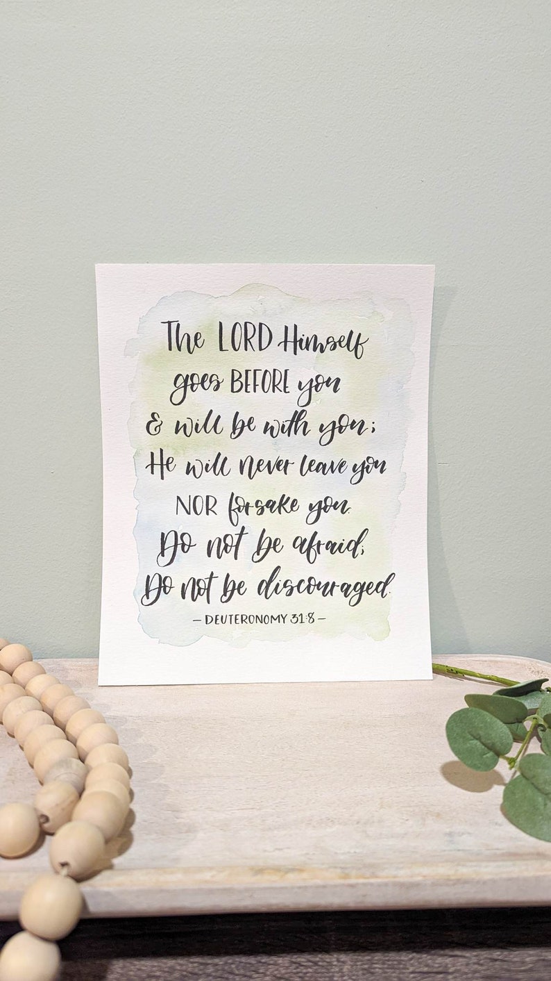 May include: A watercolor print with a light blue, green, and yellow background. The print features a handwritten quote from Deuteronomy 31:8, which reads: "The LORD Himself goes BEFORE you & will be with you; He will never leave you NOR forsake you Do not be afraid Do not be discouraged - DEUTERONOMY 31:8 -"