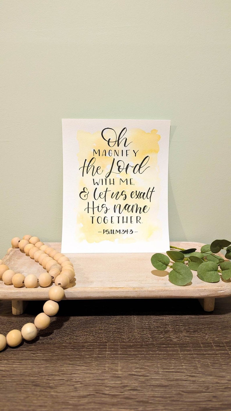 May include: A watercolor print with a yellow background and black calligraphy that reads "Oh Magnify the Lord with me & let us exalt His name together. -Psalm 34:3-"