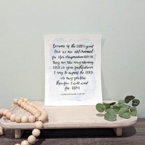 May include: A watercolor print with a blue and white background featuring a bible verse from Lamentations 3:22-24. The verse reads "Because of the LORD'S great love we are not consumed, for His compassions never fail. They are new every morning. Great is your faithfulness. I say to myself, the LORD is my portion. Therefore I will wait for HIM."