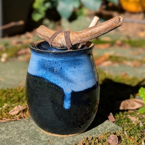Peut inclure: Un pot en céramique fait à la main avec un glaçage bicolore noir et bleu. Un morceau de bois flotté est fixé au sommet avec une lanière en cuir. Le pot est posé sur une surface en pierre moussue, avec un arrière-plan flou de verdure.