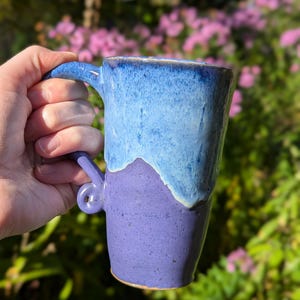 May include: A handcrafted ceramic mug with a two-tone design. The top half is a gradient of light to dark blue, while the bottom half is a deep purple. The mug has a curved handle and a textured surface.