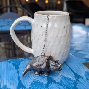 May include: A white ceramic mug with a textured surface and a large handle. A dinosaur-shaped tea infuser hangs from the rim of the mug. The infuser is dark brown with bronze accents. The mug sits on a blue patterned surface.