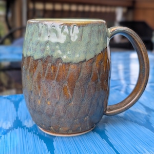 May include: A handmade ceramic mug with a textured surface. The mug features a color gradient, with a teal green top, a brown middle section, and a darker blue-brown base. The handle is a matching brown color. The mug is designed for hot beverages.