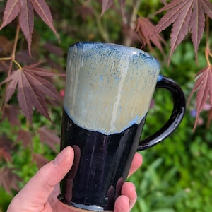 May include: A handcrafted ceramic mug with a distinctive design. The mug has a black base, a tan mid-section, and a blue-glazed rim. The handle is black. The mug is held in a hand, with a blurred background of green foliage and red leaves.