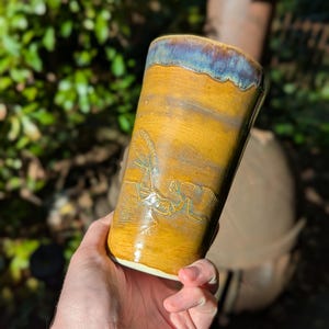 May include: A cylindrical ceramic tumbler featuring a mustard yellow glaze with a dark blue and purple rim. The exterior surface displays a relief carving of a praying mantis insect, showcasing detailed wings and legs in a light tan tone.
