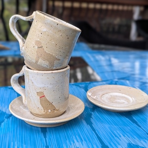 May include: Two beige ceramic mugs with a dog silhouette design, stacked. One mug sits on a matching saucer. The mugs have a speckled finish and heart-shaped handles. A second saucer is also visible.