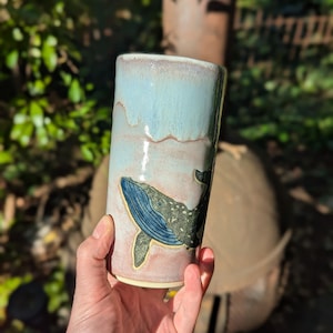 May include: A cylindrical ceramic tumbler featuring a glossy glaze with a light blue upper section and a muted pink lower section. A detailed dark blue and green whale illustration is embossed on the side of the vessel, showing its fins and tail.