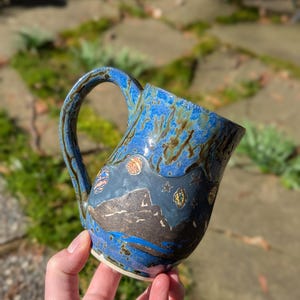 Könnte beinhalten: Eine handgefertigte Keramiktasse mit blauer Glasur und Landschaftsdesign. Die Tasse hat einen dunkelblauen Boden mit einem Gebirge und himmlischen Akzenten. Der Griff ist dunkler blau mit braunen Streifen.