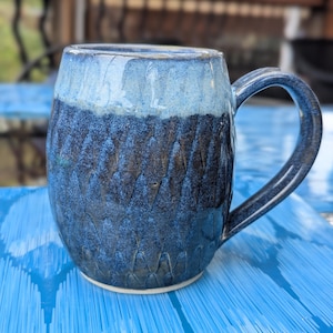 Pode incluir: Uma caneca de cerâmica artesanal com um exterior azul escuro texturizado e uma borda azul mais clara. A caneca tem uma alça grande e uma base branca, ideal para café ou chá. A caneca tem cerca de 10 cm de altura.