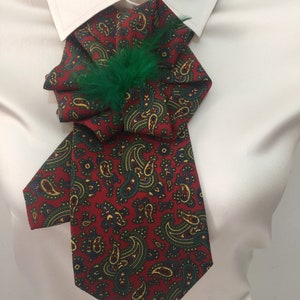 May include: A red and green paisley patterned bow tie with a green feather accent. The bow tie is tied around a white collared shirt.