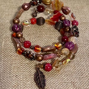 May include: A multi-strand bracelet with a variety of beads in shades of red, purple, gold, and brown. The bracelet includes round, oval, and faceted beads, with a copper-colored feather charm.