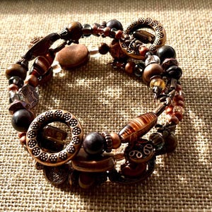 May include: A multi-strand bracelet featuring a mix of beads in shades of brown, copper, and clear. The bracelet includes various shapes and sizes of beads, including round, cylindrical, and patterned elements. The clasp is a decorative copper-toned ring.