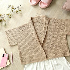 Può includere: Un accappatoio beige a maniche corte e aperto sul davanti è appoggiato su una superficie chiara con venature del legno, accanto a pantaloncini bianchi. La scena comprende un telefono rosa, un taccuino, una penna, una tazza di caffè e un piatto di cibo. Sono visibili anche pantofole rosa e un cuscino.