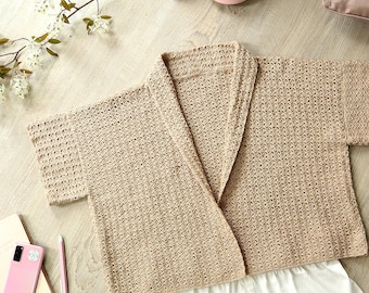 Puede incluir: Una bata beige de manga corta y abierta por delante está colocada sobre una superficie clara con vetas de madera, junto a unos pantalones cortos blancos. La escena incluye un teléfono rosa, un cuaderno, un bolígrafo, una taza de café y un plato de comida. También se ven zapatillas rosas y una almohada.