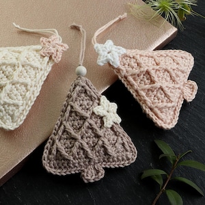 May include: Three crocheted Christmas tree ornaments in beige, pink, and brown with white crocheted snowflakes. The ornaments are hanging from a string.