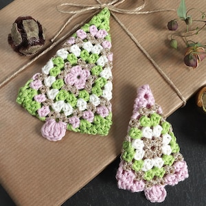 Puede incluir: Dos adornos de árbol de Navidad de ganchillo en verde, blanco, rosa y dorado, sobre un regalo envuelto en papel marrón. Los árboles tienen un aspecto hecho a mano, con una pequeña base rosa. También se ven una piña y bolas doradas.
