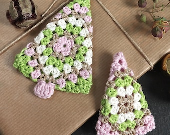 May include: Two crocheted Christmas tree ornaments in green, white, pink, and gold, resting on a brown paper-wrapped gift. The trees have a textured, handmade appearance, with a small pink base. A pine cone and gold baubles are also visible.