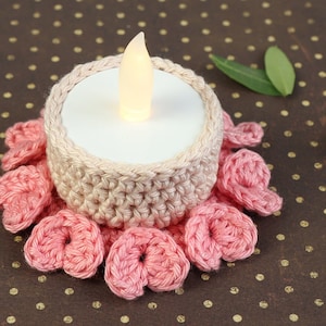 May include: A crocheted candle holder with a pink and beige color scheme. The holder is decorated with a row of pink crocheted flowers around the base. A small white tea light candle is placed inside the holder.