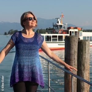 May include: A woman wearing a blue and purple crochet top with a loose, open weave. She is standing on a dock with a ferry boat in the background.