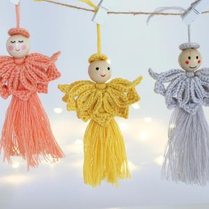 Può includere: Tre decorazioni ad angelo in crochet con teste in legno e nappe di filo. Gli angeli sono rosa, gialli e grigi.