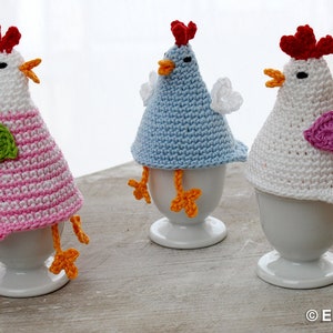 May include: Three crocheted chicken egg cozies in pink, blue, and white. Each cozie has a small heart detail and sits on a white ceramic egg cup.